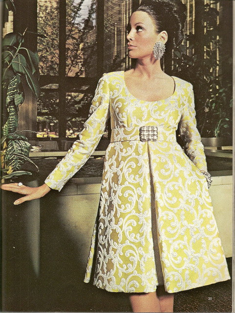 A brunette woman wears a white and gold brocade empire waist mini dress with long sleeves and large diamond earrings