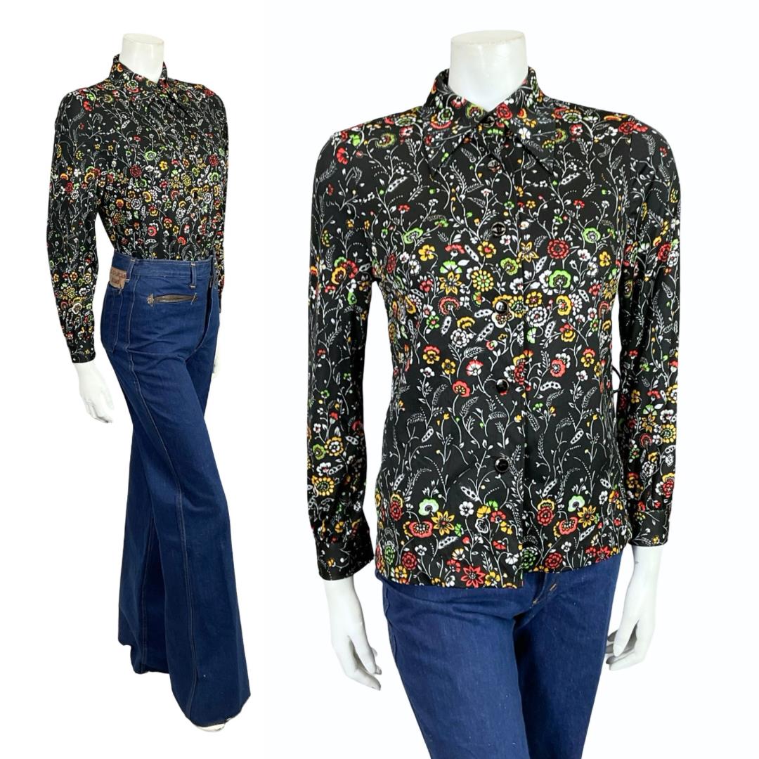 Mannequin wears a black white green and red floral printed 60s 70s dagger collar shirt with blue flared jeans. 